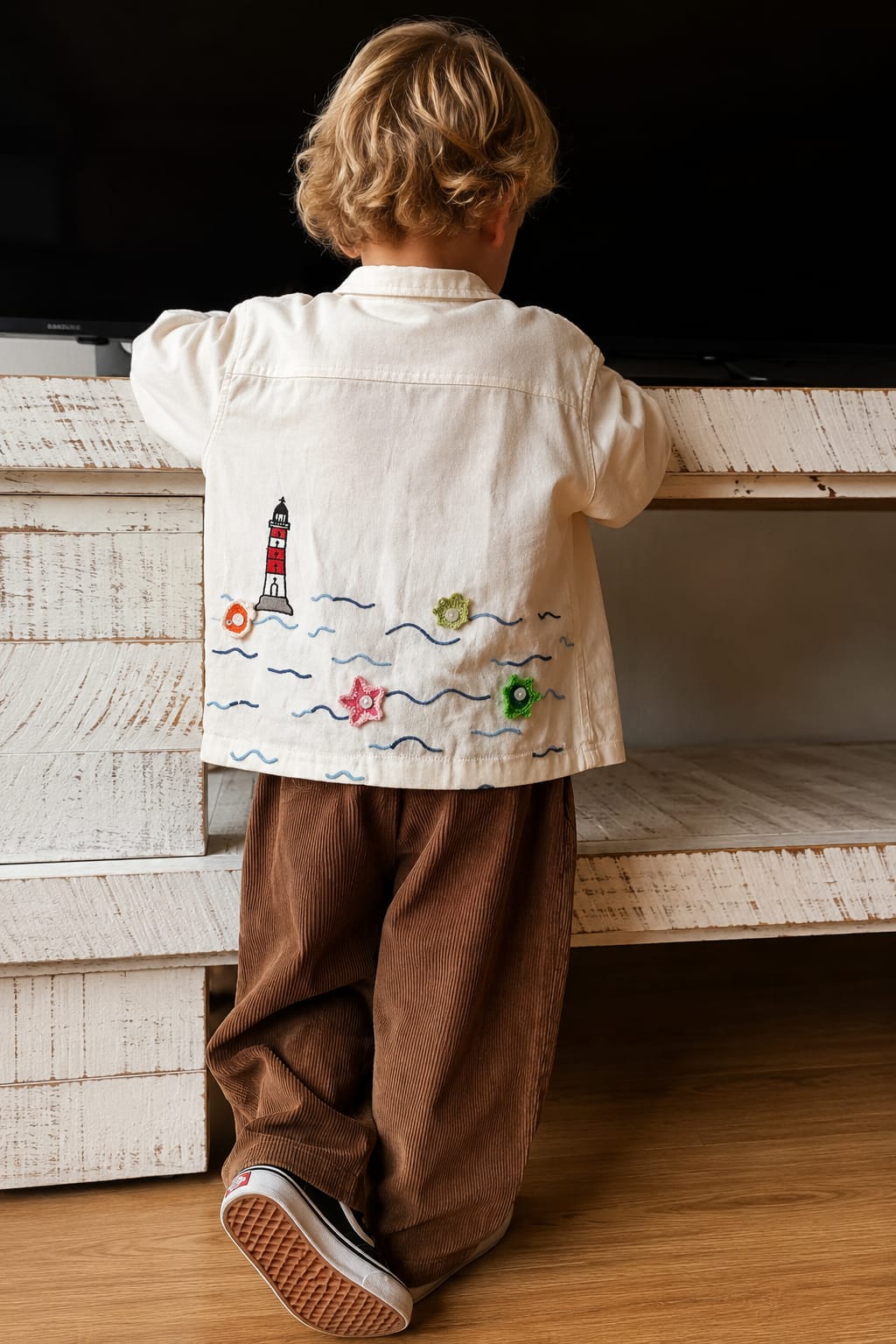 A child viewed from behind wearing a cream-colored shirt with a colorful lighthouse and wave embroidery on the back, paired with brown pants, standing in front of a white wooden shelf.