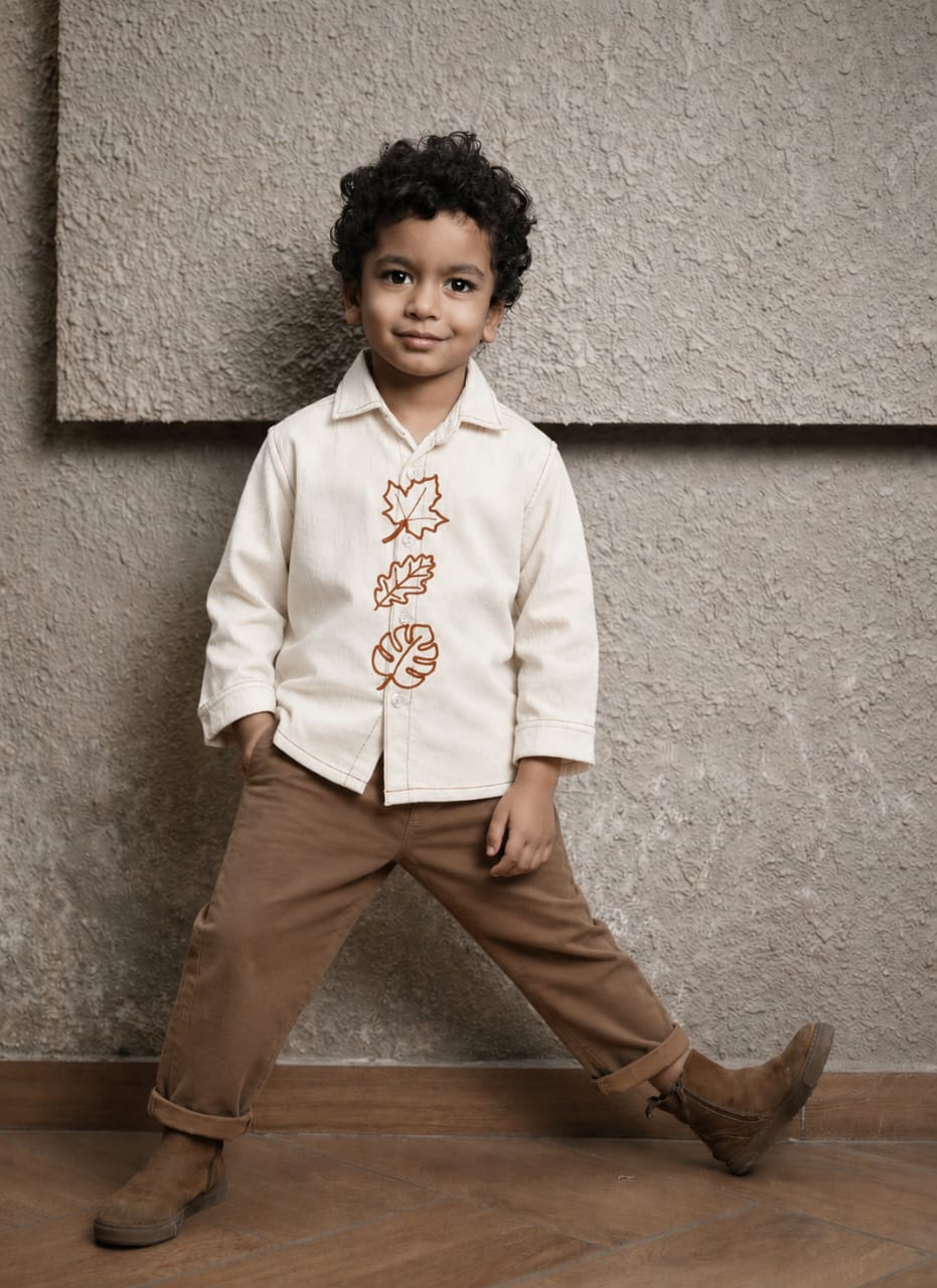 A young boy wearing a cream-colored long-sleeve button-up shirt with brown embroidered leaf motifs down the front, paired with brown pants and brown boots, posing against a textured beige wall.