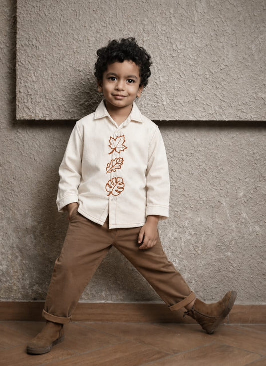 A young boy wearing a cream-colored long-sleeve button-up shirt with brown embroidered leaf motifs down the front, paired with brown pants and brown boots, posing against a textured beige wall.