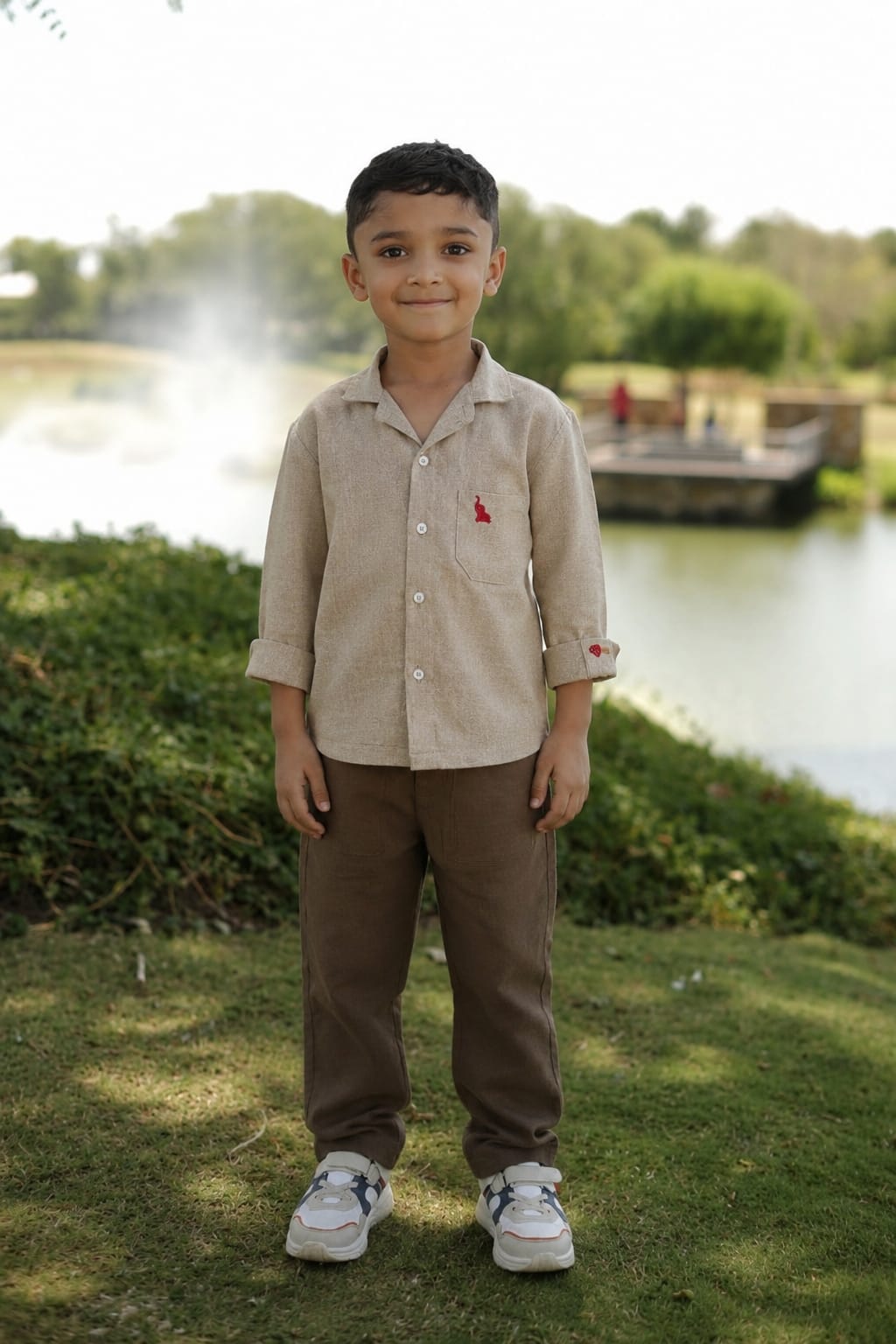 A young boy standing outdoors on grass near a lake, wearing a light beige long-sleeve button-up shirt with a small red embroidered logo on the chest, brown pants, and white sneakers.