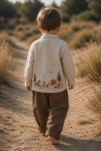 A toddler viewed from behind walking barefoot on a dirt path in a dry outdoor landscape, wearing a cream-colored long-sleeve shirt with a forest cabin embroidery on the back and brown pants.