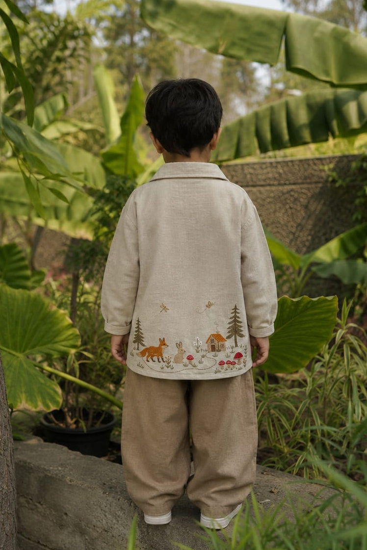 A child viewed from behind wearing a light beige embroidered jacket and matching pants, standing outdoors in a lush garden with green banana leaves and plants.