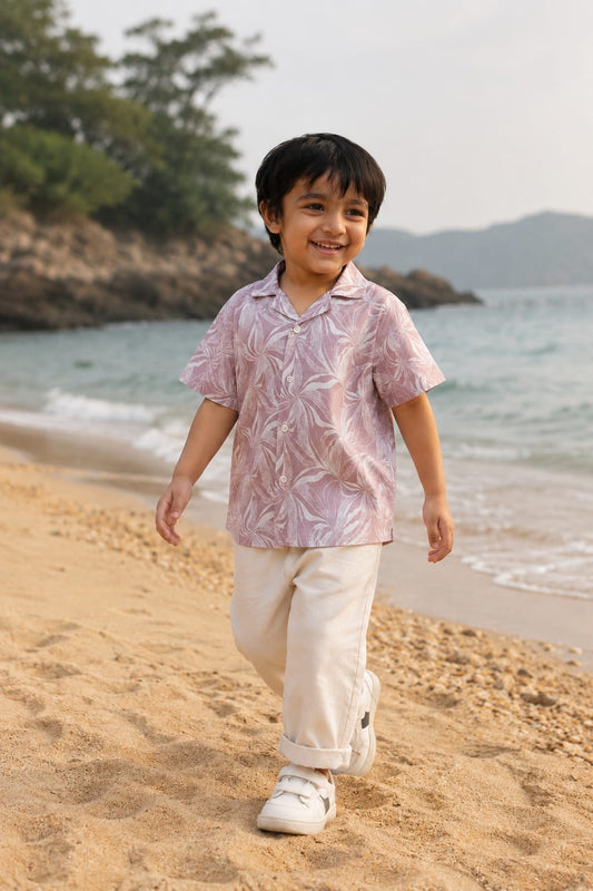 A young boy walking on a sandy beach wearing a light pink short-sleeve button-up shirt with a white tropical leaf pattern, cream pants, and white sneakers.
