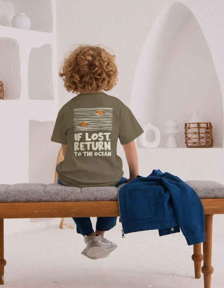 Child wearing a t-shirt with 'If Lost, Return to the Ocean' text in a minimalistic room.