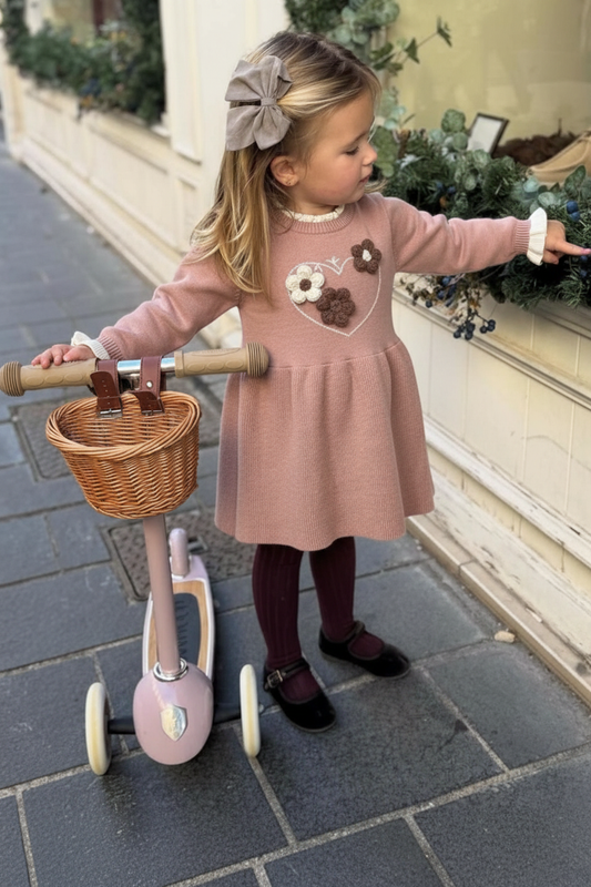 Child in a pink dress with floral design standing next to a tricycle on a sidewalk.